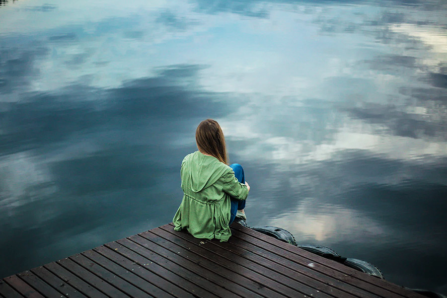 young lady on their own sat at the end of a jetty surrounded my water looking sad