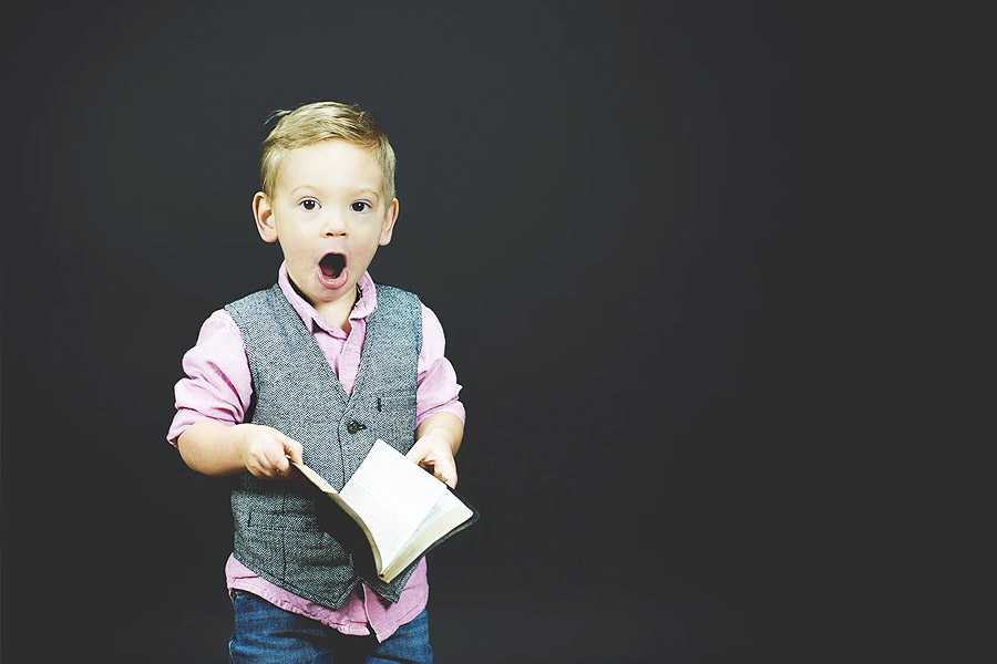 young boy on a grey background looking shocked reading a book